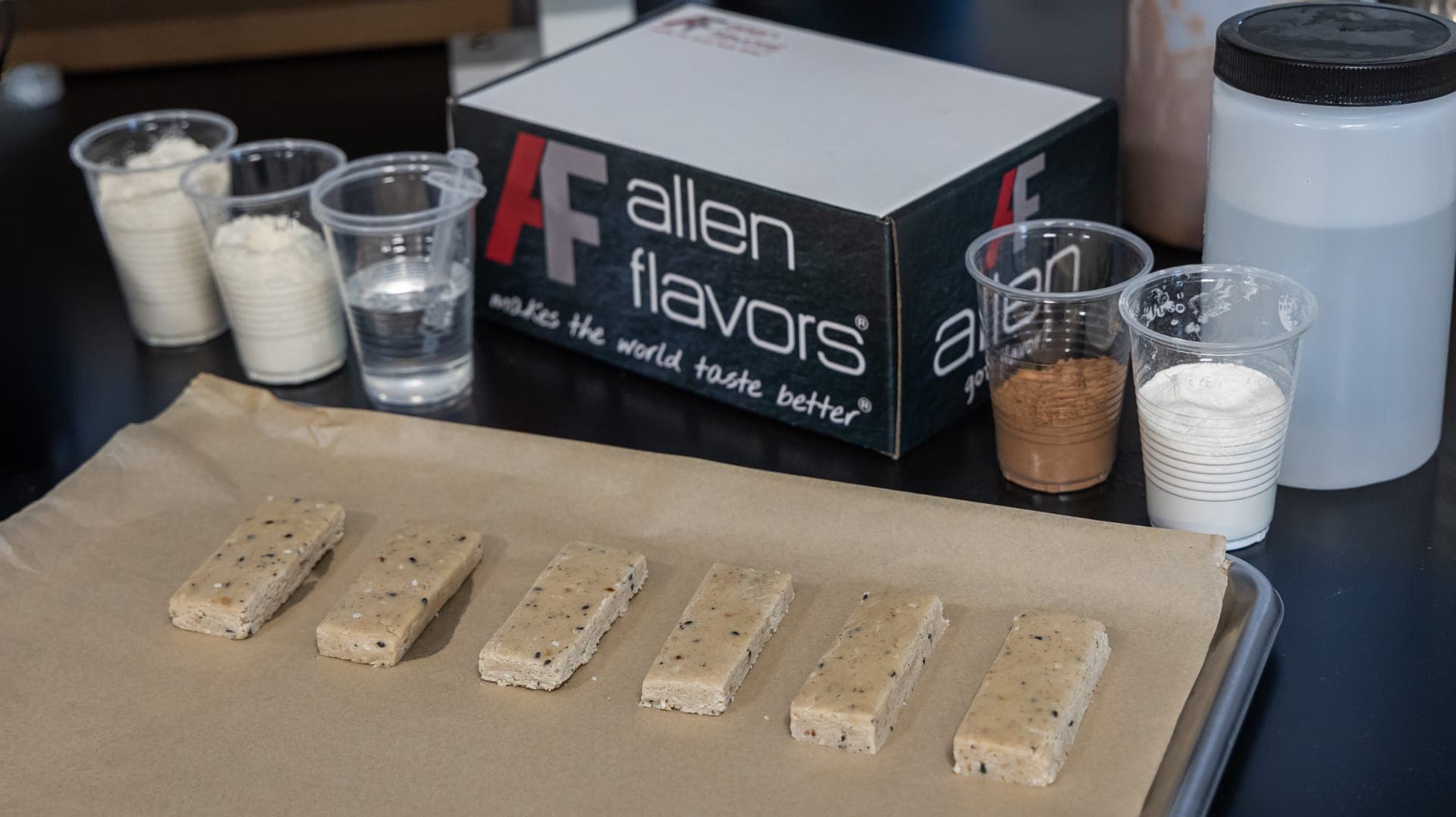 Six bar samples on parchment paper beside cups of dry ingredients and an Allen Flavors box