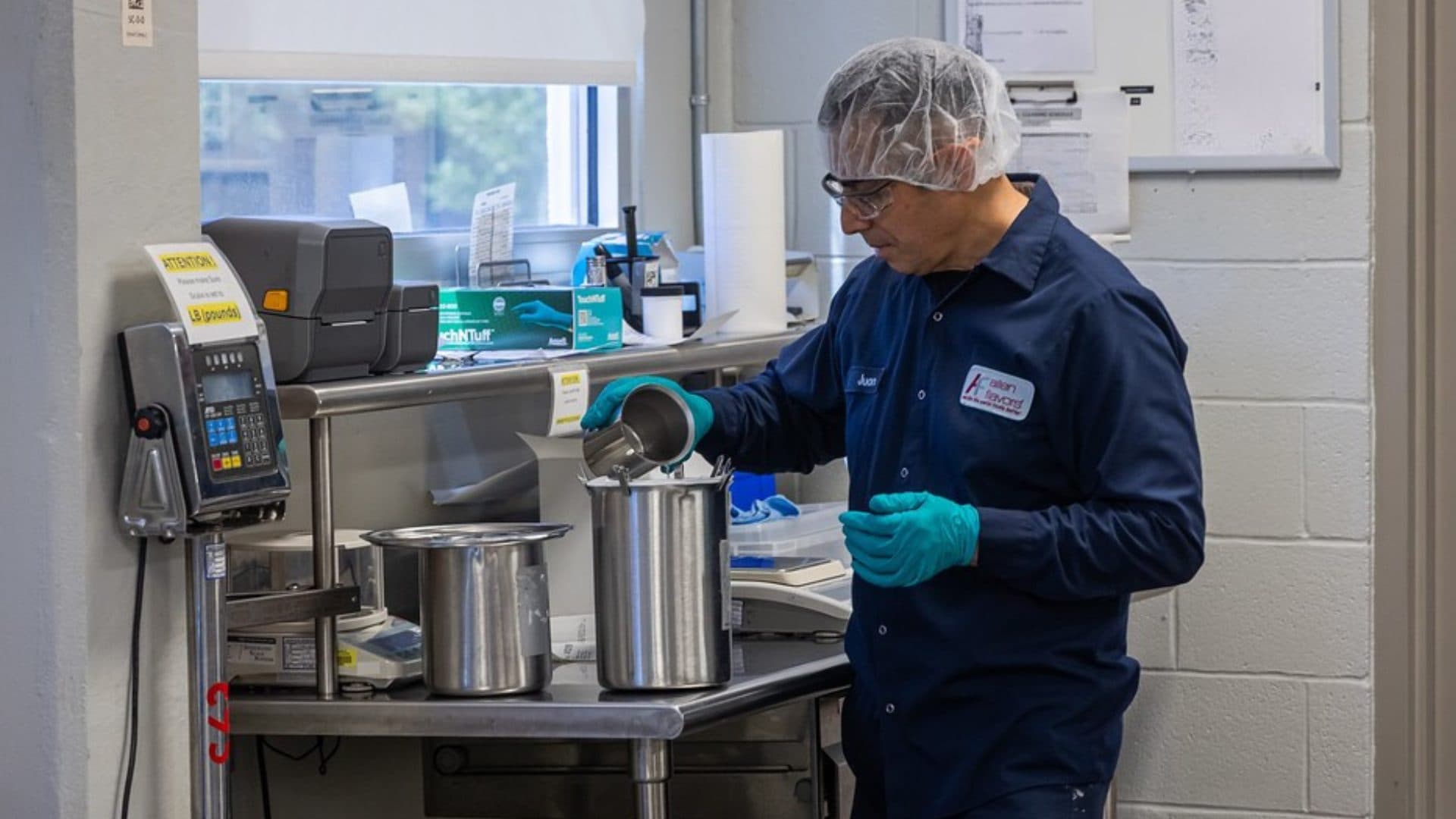 Production team member in a hairnet and safety glasses pouring an ingredient into a stainless container at a scale station