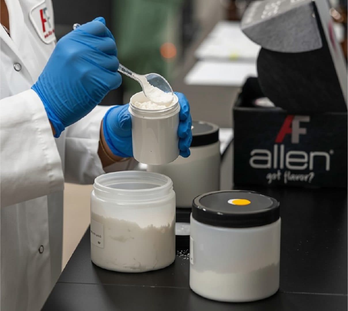Gloved hands scooping a white powder ingredient into a container on the lab bench