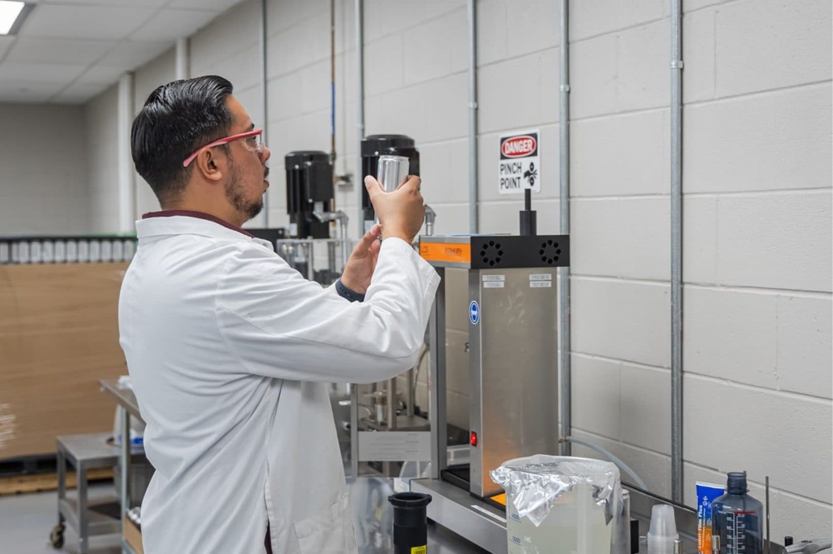 Team member in a lab coat and safety glasses checking a small container beside production equipment