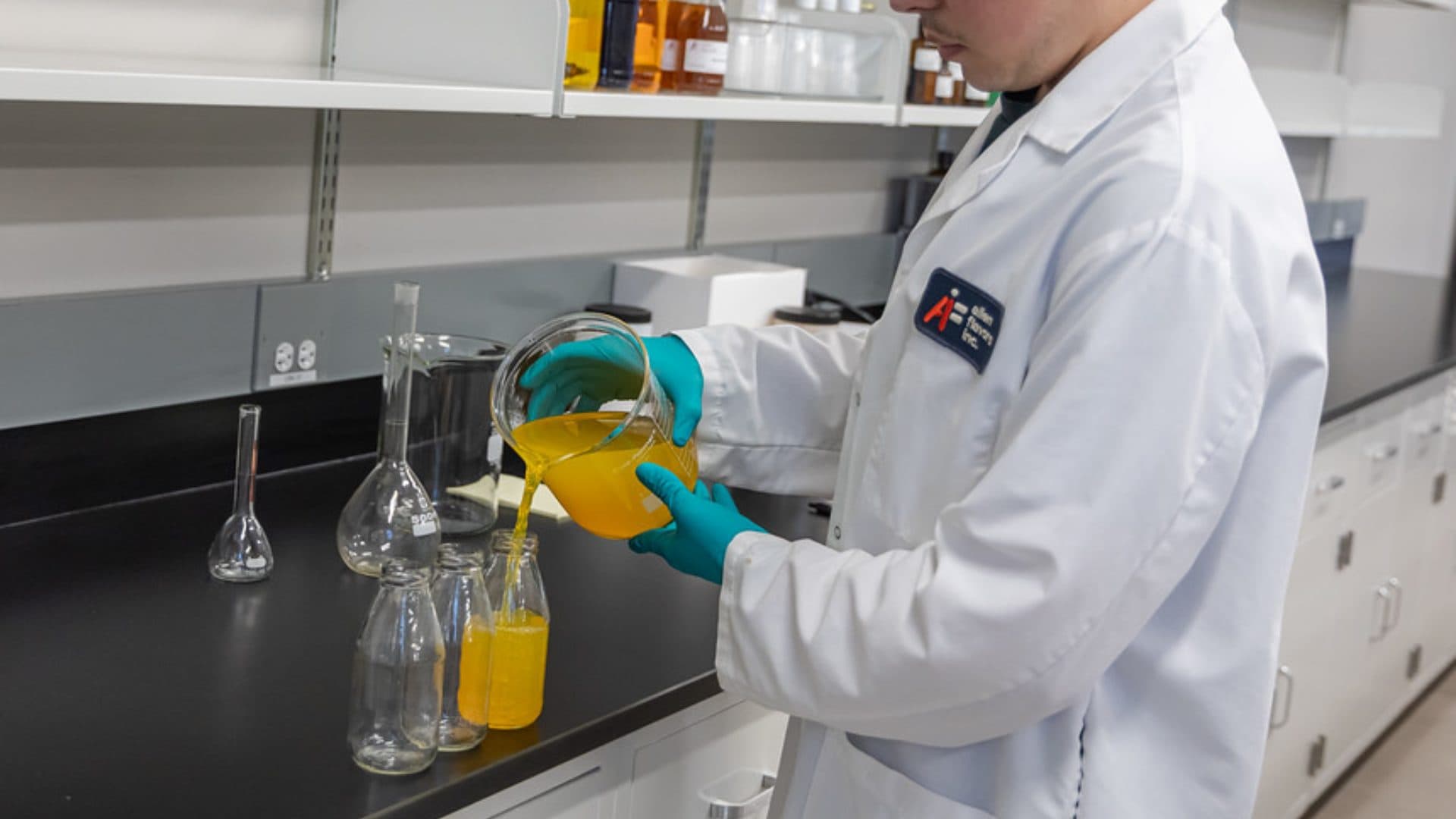 Product developer in an Allen Flavors lab coat pouring a yellow beverage sample from a beaker into glass bottles